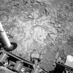 NASA’s Curiosity Achieves Drilling Milestone on Nevado Sajama A grayscale photo from the Martian surface shows uneven terrain of mostly flat, fractured rock on the ground ahead of the rover. A small drill hole and its surrounding dust is visible near the center of the image. Parts of the rover are visible at the bottom and left sides of the frame, including part of the rover arm and its joint along the left side of the image.