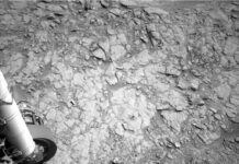 NASA’s Curiosity Achieves Drilling Milestone on Nevado Sajama A grayscale photo from the Martian surface shows uneven terrain of mostly flat, fractured rock on the ground ahead of the rover. A small drill hole and its surrounding dust is visible near the center of the image. Parts of the rover are visible at the bottom and left sides of the frame, including part of the rover arm and its joint along the left side of the image.