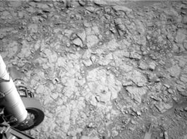 NASA’s Curiosity Achieves Drilling Milestone on Nevado Sajama A grayscale photo from the Martian surface shows uneven terrain of mostly flat, fractured rock on the ground ahead of the rover. A small drill hole and its surrounding dust is visible near the center of the image. Parts of the rover are visible at the bottom and left sides of the frame, including part of the rover arm and its joint along the left side of the image.