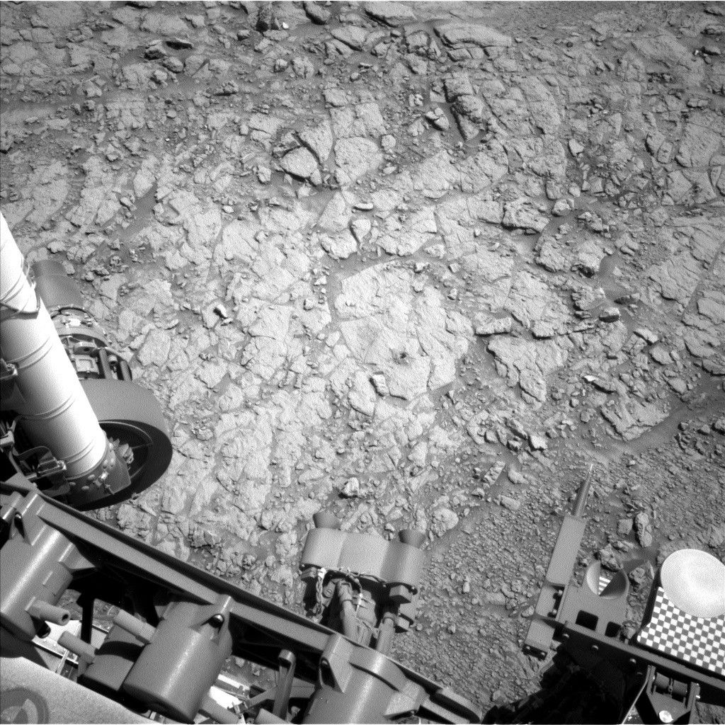 A grayscale photo from the Martian surface shows uneven terrain of mostly flat, fractured rock on the ground ahead of the rover. A small drill hole and its surrounding dust is visible near the center of the image. Parts of the rover are visible at the bottom and left sides of the frame, including part of the rover arm and its joint along the left side of the image.