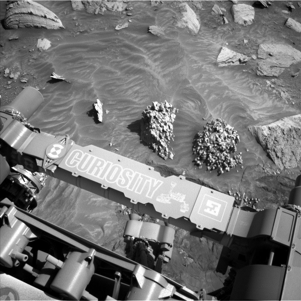 https mars.nasa .gov msl raw images proj msl redops ods surface sol 04478 opgs edr ncam nlb 7950309.jpeg A grayscale photograph of the Martian surface by the Curiosity rover captures medium gray soil with exposed small to medium sized rocks scattered around. Two medium sized rocks just in front of the rover stand out – unlike the smooth or sharp-edged ones around them, they look very rough-textured and bumpy, like a large clump of granola. The bottom of the frame shows parts of the rover, running from the middle left edge to the lower right corner of the image, including part of its robotic arm which carries a nameplate imprinted with “Curiosity” outlined in white, all-capital letters, and to the right of that a line drawing of the rover.
