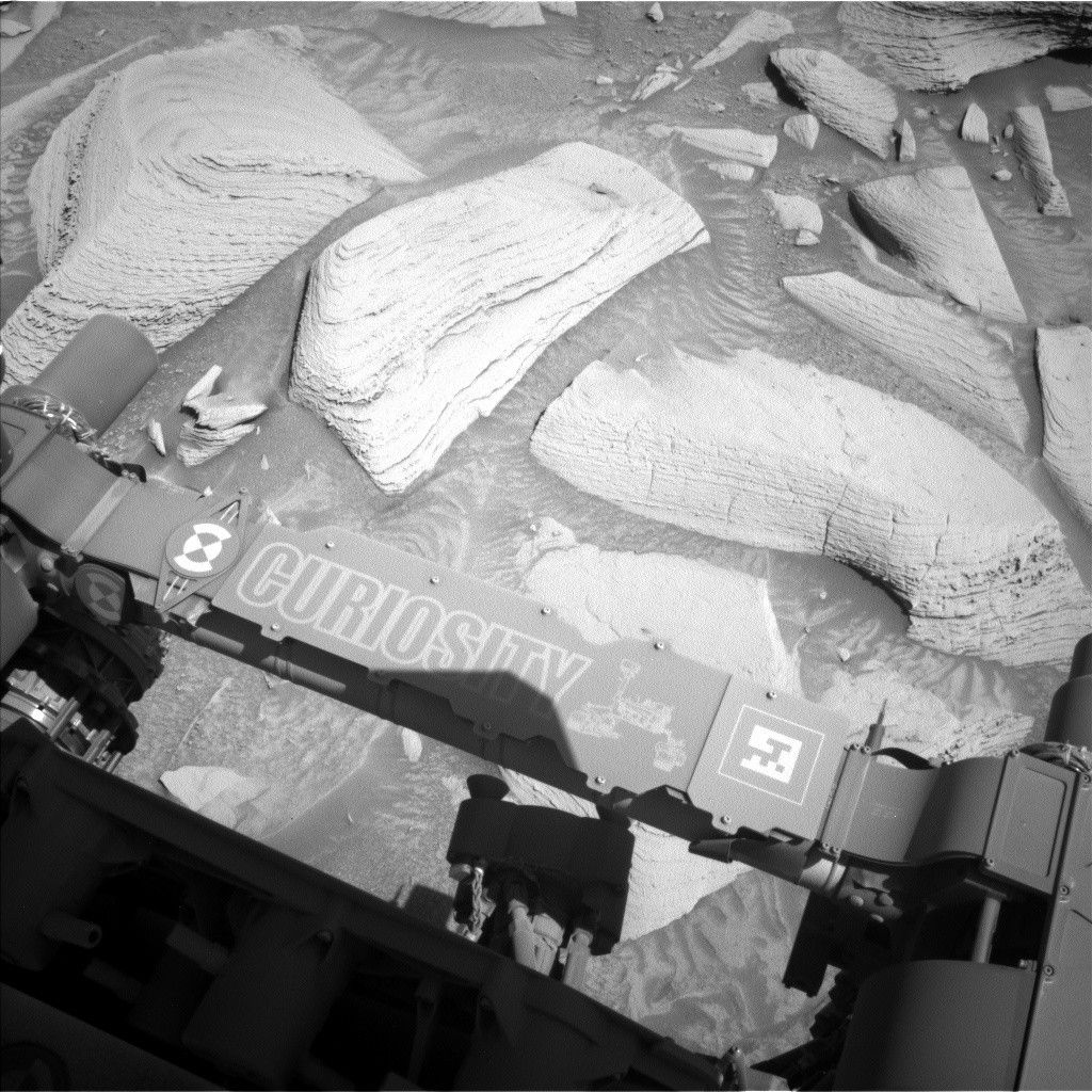 https mars.nasa .gov msl raw images proj msl redops ods surface sol 04483 opgs edr ncam nlb 7954724.jpeg A grayscale photograph of the Martian surface by the Curiosity rover captures medium gray soil next to the rover with exposed medium-sized, lighter-colored rocks scattered around. Most of the rocks appear layered, with dividing lines making them look like features drawn on a topographic map. The bottom of the frame shows parts of the rover, running from the middle left edge to the lower right corner of the image, including part of its robotic arm which carries a nameplate imprinted with “Curiosity” outlined in white, all-capital letters, and to the right of that a line drawing of the rover.
