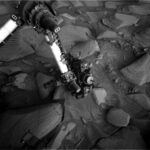 Navigating Rough Terrain: Sols 4488-4490 South of Texoli Butte A grayscale photo from the Martian surface shows the Curiosity rover’s robotic arm reaching down to investigate a targeted rock. The surrounding terrain is very rocky and uneven, with stones of varied shapes and sizes pushing up out of the ground, everything a similar dark shade of gray. The soil between the rocks appears wavy in many places, as if sculpted by wind or water, and most of the rocks look like they have formed from horizontal layers stacking on one another, in the relatively short elevation they rise from the surface, looking like 3D versions of hills indicated on a topographic map. The rover’s arm extends down to the center of the image from the upper left corner of the frame where, at a pivot point, it bends at roughly 90-degree angle toward the left side of the image and out of the frame. The arm itself is bright-toned, nearly white, with a metallic sheen; the pivot point and the boxy suite of instruments at the end of the arm near the ground are mainly dark gray or black, with multiple knobby protrusions.
