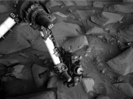 Navigating Rough Terrain: Sols 4488-4490 South of Texoli Butte A grayscale photo from the Martian surface shows the Curiosity rover’s robotic arm reaching down to investigate a targeted rock. The surrounding terrain is very rocky and uneven, with stones of varied shapes and sizes pushing up out of the ground, everything a similar dark shade of gray. The soil between the rocks appears wavy in many places, as if sculpted by wind or water, and most of the rocks look like they have formed from horizontal layers stacking on one another, in the relatively short elevation they rise from the surface, looking like 3D versions of hills indicated on a topographic map. The rover’s arm extends down to the center of the image from the upper left corner of the frame where, at a pivot point, it bends at roughly 90-degree angle toward the left side of the image and out of the frame. The arm itself is bright-toned, nearly white, with a metallic sheen; the pivot point and the boxy suite of instruments at the end of the arm near the ground are mainly dark gray or black, with multiple knobby protrusions.
