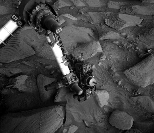 Navigating Rough Terrain: Sols 4488-4490 South of Texoli Butte A grayscale photo from the Martian surface shows the Curiosity rover’s robotic arm reaching down to investigate a targeted rock. The surrounding terrain is very rocky and uneven, with stones of varied shapes and sizes pushing up out of the ground, everything a similar dark shade of gray. The soil between the rocks appears wavy in many places, as if sculpted by wind or water, and most of the rocks look like they have formed from horizontal layers stacking on one another, in the relatively short elevation they rise from the surface, looking like 3D versions of hills indicated on a topographic map. The rover’s arm extends down to the center of the image from the upper left corner of the frame where, at a pivot point, it bends at roughly 90-degree angle toward the left side of the image and out of the frame. The arm itself is bright-toned, nearly white, with a metallic sheen; the pivot point and the boxy suite of instruments at the end of the arm near the ground are mainly dark gray or black, with multiple knobby protrusions.