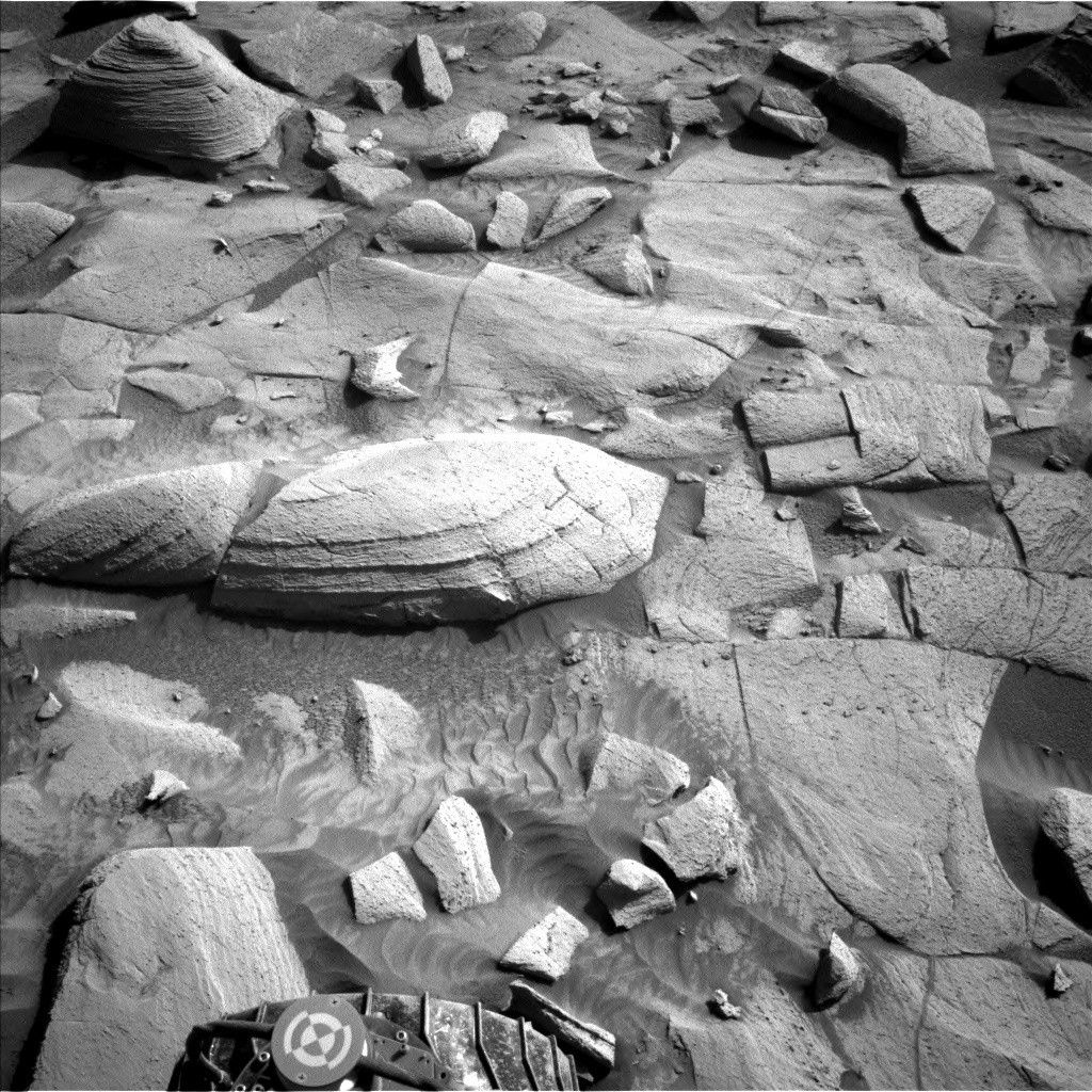 A grayscale photograph of the Martian surface from the Curiosity rover captures medium gray soil in front of the rover, wavy in some areas, as if sculpted by wind or water, with rocks everywhere protruding up through it. They are lighter-toned than the ground, some rounded, others looking like cracked, smooth plates. One larger, rounded, light-colored rock stands out, in the center of the image. A part of the rover is visible in the frame, at bottom center.