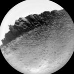 NASA Science: Sols 4495-4497 – Yawning, Perching, Rolling A circular grayscale image from the Martian surface is skewed slightly left, so the horizon line runs from about the 8 o’clock position on a clock face to about 2 o’clock. Gritty, gravelly terrain in the foreground leads to a spiky, ragged outcrop with vertical ridges and grooves rising on the horizon.