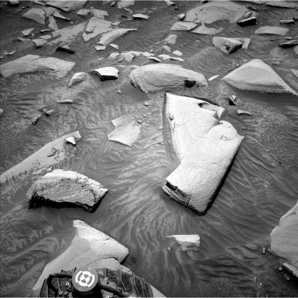 https mars.nasa .gov msl raw images proj msl redops ods surface sol 04494 opgs edr ncam nlb 7964497.jpeg A grayscale photograph of the Martian surface from the Curiosity rover captures medium gray soil in front of the rover, wavy in most areas, as if sculpted by wind or water, with rocks everywhere protruding up through it. They are lighter-toned than the ground, some rounded, others looking like cracked, smooth plates. One large, jagged, light-colored rock stands out, in the center of the image. A part of the rover is visible in the frame, at bottom center.