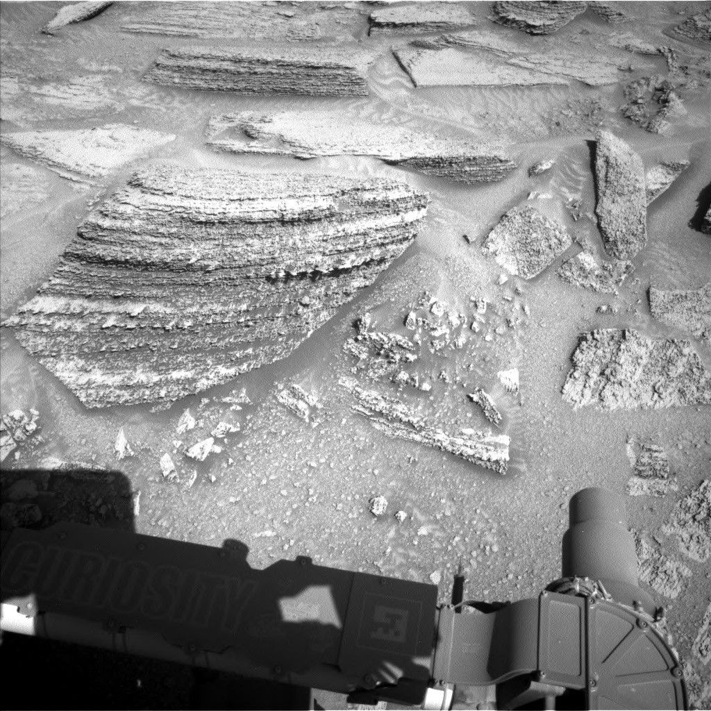 A grayscale close-up photograph of the Martian surface from the Curiosity rover shows a mix of small to large medium-gray rocks exposed above coarse soil. The rocks are various polygonal shapes with mostly sharp edges; the largest stands out in particular on the upper left side of the image, composed of numerous rough layers viewed edge-on. Portions of the rover are also visible in the frame, mainly Curiosity’s robotic arm, in a shadow at the bottom of the frame. The rover’s name is printed on the arm in all uppercase letters, next to a silhouette image of the rover.