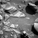 NASA Science Explores Sols 4525-4526: Journey South Near Texoli A grayscale photograph of the Martian surface from the Curiosity rover captures medium-gray, coarse soil in front of the rover, with rocks everywhere protruding up through it. They are lighter-toned than the ground, some rounded, others looking like cracked, smooth plates. The largest rocks look like they’re built up from stacked layers. A part of the rover is visible in the frame, at bottom center, including one of its wheels.