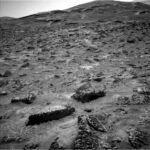 Sols 4527-4528: NASA Discovers ‘Boxwork’ Formation A grayscale photograph of the Martian surface from the Curiosity rover shows a field of rough and jagged rocks covering the surface from the foreground extending to hills in the background, all medium to dark gray. The hills appear smoother overall, but have horizontal folds on their sides leading up to their peaks.