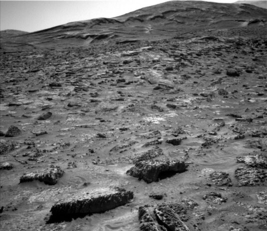 Sols 4527-4528: NASA Discovers ‘Boxwork’ Formation A grayscale photograph of the Martian surface from the Curiosity rover shows a field of rough and jagged rocks covering the surface from the foreground extending to hills in the background, all medium to dark gray. The hills appear smoother overall, but have horizontal folds on their sides leading up to their peaks.