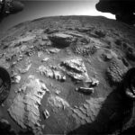 Mars Exploration: Discovering Honeycomb and Waffle Patterns An extreme wide-angle photo of the Martian surface in front of the Curiosity rover shows extremely rocky, rough terrain, with flat rocks in the foreground showing apparent honeycomb-shaped patterns on their exposed faces, and a larger rock at the center of the image, looking like an offset stack of waffles. Portions of the rover are visible in each corner of the frame, with two wheels visible, but shadowed, in the lower left and right corners.