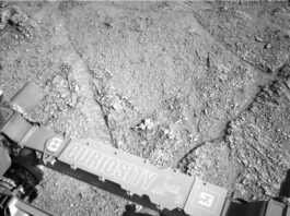 After Busy Weekend, Sols 4539-4540 Mark NASA Science Return A grayscale photograph of Martian terrain in front of the Curiosity rover shows flat, rough ground covered in gravel, with large, polygonal sections of rock on the surface separated by shallow gouges in the soil. The bottom of the frame shows parts of the rover, running from the middle left edge to the lower right corner of the image, including part of its robotic arm which carries a nameplate imprinted with “Curiosity” outlined in white, all-capital letters, and to the right of that a line drawing of the rover.