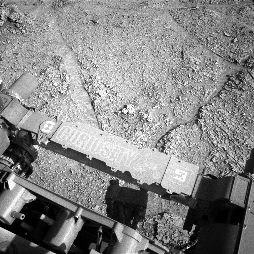 https mars.nasa .gov msl raw images proj msl redops ods surface sol 04537 opgs edr ncam nlb 8002704.jpeg A grayscale photograph of Martian terrain in front of the Curiosity rover shows flat, rough ground covered in gravel, with large, polygonal sections of rock on the surface separated by shallow gouges in the soil. The bottom of the frame shows parts of the rover, running from the middle left edge to the lower right corner of the image, including part of its robotic arm which carries a nameplate imprinted with “Curiosity” outlined in white, all-capital letters, and to the right of that a line drawing of the rover.