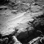 NASA Investigates: True Boxwork or Merely Box-Like Structure? A grayscale photograph of Martian terrain in front of the Curiosity rover shows a ledge of flat, rough ground that appears to drop off just in front of the rover. Portions of the rover are visible at the bottom of the frame, including a wheel in the lower-right corner. An area of the ground ahead is smoother than the rest, looking like small, wavy sand dunes. Many of the small to medium sized rocks surrounding this patch have trails of sandy soil running off to their right, as if wind or water had flowed from left to right and sculpted these pointed tails of runoff.