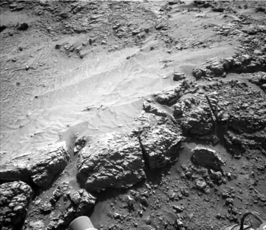 Days 4543-4545: Departing Ridge to Explore New Terrains A grayscale photograph of Martian terrain in front of the Curiosity rover shows a ledge of flat, rough ground that appears to drop off just in front of the rover. Portions of the rover are visible at the bottom of the frame, including a wheel in the lower-right corner. An area of the ground ahead is smoother than the rest, looking like small, wavy sand dunes. Many of the small to medium sized rocks surrounding this patch have trails of sandy soil running off to their right, as if wind or water had flowed from left to right and sculpted these pointed tails of runoff.