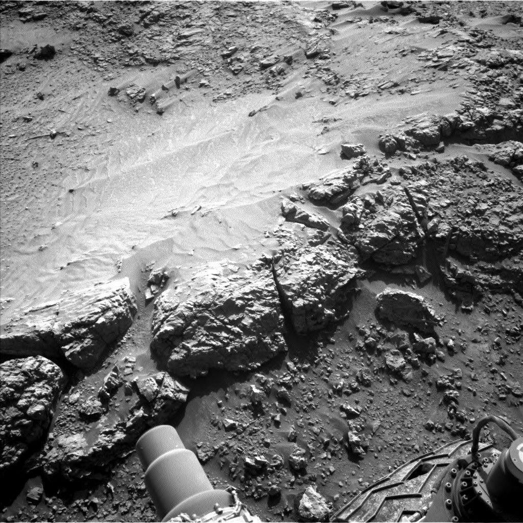 https mars.nasa .gov msl raw images proj msl redops ods surface sol 04541 opgs edr ncam nlb 8006247.jpeg A grayscale photograph of Martian terrain in front of the Curiosity rover shows a ledge of flat, rough ground that appears to drop off just in front of the rover. Portions of the rover are visible at the bottom of the frame, including a wheel in the lower-right corner. An area of the ground ahead is smoother than the rest, looking like small, wavy sand dunes. Many of the small to medium sized rocks surrounding this patch have trails of sandy soil running off to their right, as if wind or water had flowed from left to right and sculpted these pointed tails of runoff.