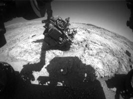 Attempting Again: Sols 4554–4555 NASA Science Update A grayscale wide-angle view of the Martian surface (creating an exaggerated, curved horizon) shows very rough terrain in front of the Curiosity rover. Light gray, gravel-covered rocks extend to the horizon, separated by gouges in some areas. Reaching down from the top of the frame is Curiosity’s robotic arm, with the turret at its end holding science instruments reaching toward the ground. The rover’s shadow, including its body and robotic arm, darkens the bottom third of the image.