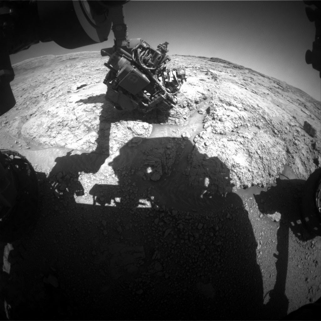 A grayscale wide-angle view of the Martian surface (creating an exaggerated, curved horizon) shows very rough terrain in front of the Curiosity rover. Light gray, gravel-covered rocks extend to the horizon, separated by gouges in some areas. Reaching down from the top of the frame is Curiosity’s robotic arm, with the turret at its end holding science instruments reaching toward the ground. The rover’s shadow, including its body and robotic arm, darkens the bottom third of the image.