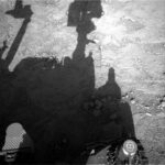 NASA Science: Daily Box Tasks on Sols 4556-4558 A grayscale photo looking down at the Martian surface, showing very rough terrain in front of the Curiosity rover. Light gray, gravel-covered rocks are in bright sunlight in about half the frame, extending up diagonally toward the upper right corner of the image. A shadow silhouette of the rover covers most of the rest of the image, with its mast head and robotic arm appearing very prominently. Portions of the rover itself are shadowed but visible at the bottom of the image.