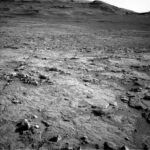 Curiosity Explores Uyuni Quad: NASA’s Latest Science Update A grayscale photograph of the Martian surface from the Curiosity rover shows a mostly flat, stony field, with rough and jagged medium-sized rocks scattered around, extending to hills in the background, all medium to dark gray. The hills appear smoother overall, with waves on the left side that look like windblown dunes.