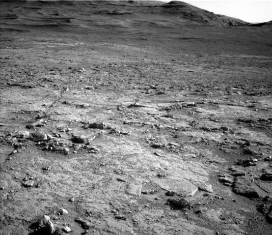 Curiosity Explores Uyuni Quad: NASA’s Latest Science Update A grayscale photograph of the Martian surface from the Curiosity rover shows a mostly flat, stony field, with rough and jagged medium-sized rocks scattered around, extending to hills in the background, all medium to dark gray. The hills appear smoother overall, with waves on the left side that look like windblown dunes.