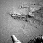 Curiosity Sols 4600-4601: Navigating the Sandy Slope – NASA Update A grayscale photo of the Martian surface shows an overhead view of a large, medium-gray rock, partially buried in sandy soil of the same color. Jagged edges and ridges of the rock stick out above the sand at various points and angles. Parts of the Curiosity rover -- a cylindrical nozzle and the edge of one wheel — are visible at the bottom of the frame.