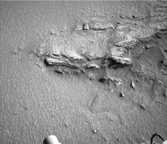Curiosity Sols 4600-4601: Navigating the Sandy Slope – NASA Update A grayscale photo of the Martian surface shows an overhead view of a large, medium-gray rock, partially buried in sandy soil of the same color. Jagged edges and ridges of the rock stick out above the sand at various points and angles. Parts of the Curiosity rover -- a cylindrical nozzle and the edge of one wheel — are visible at the bottom of the frame.