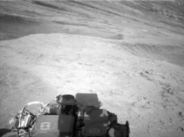 NASA’s Curiosity Explores Ridge Peaks: Sols 4602-4603 Update A grayscale photo shows Martian terrain, which appears medium gray throughout. In front of the Curiosity rover, the ground from the bottom to the middle of the frame appears relatively smooth, with a few scattered small rocks poking above the surface. In the upper part of the image, the terrain appears to slope downward from left to right, with ridges running down along that same direction. Part of the rover is visible in the lower-right corner of the image, casting a shadow on the ground.