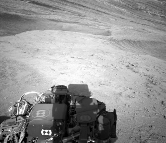NASA’s Curiosity Explores Ridge Peaks: Sols 4602-4603 Update A grayscale photo shows Martian terrain, which appears medium gray throughout. In front of the Curiosity rover, the ground from the bottom to the middle of the frame appears relatively smooth, with a few scattered small rocks poking above the surface. In the upper part of the image, the terrain appears to slope downward from left to right, with ridges running down along that same direction. Part of the rover is visible in the lower-right corner of the image, casting a shadow on the ground.