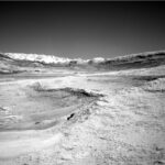 NASA Science: Curiosity Explores Deep Dip – Sols 4607-4608 A grayscale photo from the Martian surface shows a landscape of undulating but mostly smooth terrain, in light gray. A plateau on the right side of the foreground drops down toward the left about mid-frame, in a gentle curve. A ridge across the upper middle of the frame creates a V-shaped horizon line. Beyond that in the distance, filling the bottom of the V, a jagged line of rocks is brilliant white, looking like snow-capped mountains.