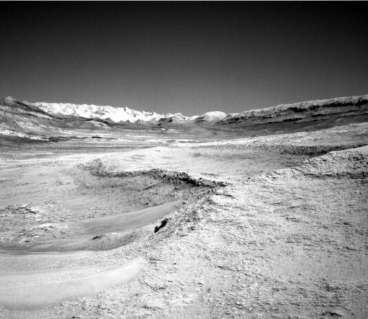 NASA Science: Curiosity Explores Deep Dip – Sols 4607-4608 A grayscale photo from the Martian surface shows a landscape of undulating but mostly smooth terrain, in light gray. A plateau on the right side of the foreground drops down toward the left about mid-frame, in a gentle curve. A ridge across the upper middle of the frame creates a V-shaped horizon line. Beyond that in the distance, filling the bottom of the V, a jagged line of rocks is brilliant white, looking like snow-capped mountains.