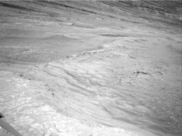 NASA Curiosity Blog: Sols 4611-4613 Scenic Overlook Exploration A grayscale photo from the Martian surface shows rocky, uneven, light-gray terrain that slopes downward, away from the rover. Parts of the rover are visible in the bottom of the frame, and the downward slope extends from the bottom of the frame toward the top of the frame, with a ridge line tracing the descent.