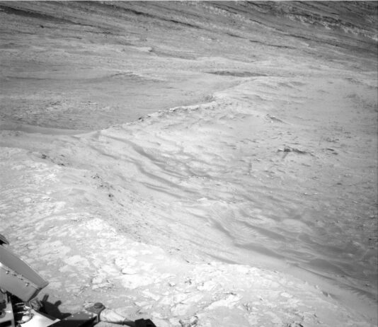 NASA Curiosity Blog: Sols 4611-4613 Scenic Overlook Exploration A grayscale photo from the Martian surface shows rocky, uneven, light-gray terrain that slopes downward, away from the rover. Parts of the rover are visible in the bottom of the frame, and the downward slope extends from the bottom of the frame toward the top of the frame, with a ridge line tracing the descent.