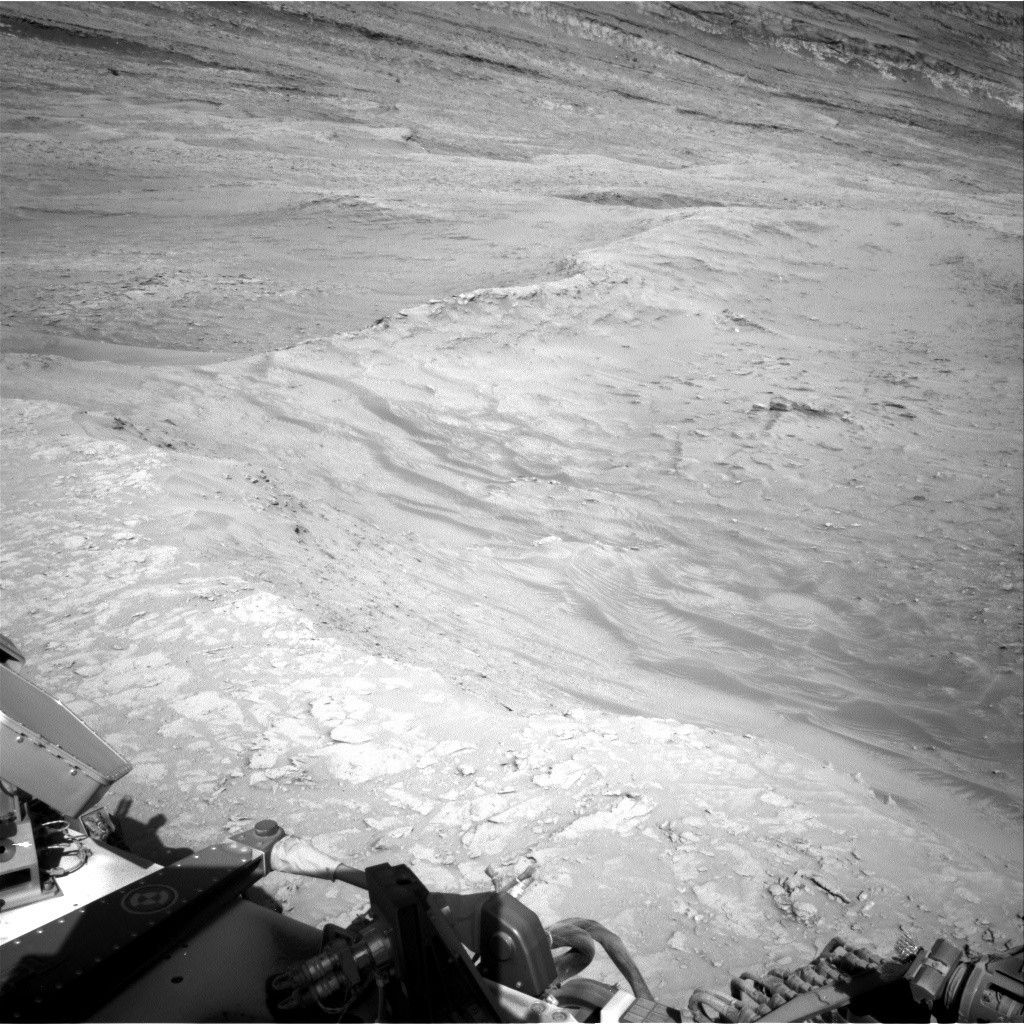 https mars.nasa .gov msl raw images proj msl redops ods surface sol 04609 opgs edr ncam nrb 8066605.jpeg A grayscale photo from the Martian surface shows rocky, uneven, light-gray terrain that slopes downward, away from the rover. Parts of the rover are visible in the bottom of the frame, and the downward slope extends from the bottom of the frame toward the top of the frame, with a ridge line tracing the descent.