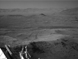 NASA’s Curiosity Explores Boxwork: Sols 4614-4615 Journey A grayscale photo from the Martian surface shows a landscape of dark gray, slightly uneven, desert-like terrain stretching off into the distance, where a line of peaks that look like sand dunes create a hazy horizon line. Part of the rover is dimly visible in the bottom of the frame, and a pair of wheel tracks, darker than the surrounding ground, extend backward from there, scratched into the soil at the center of the frame.