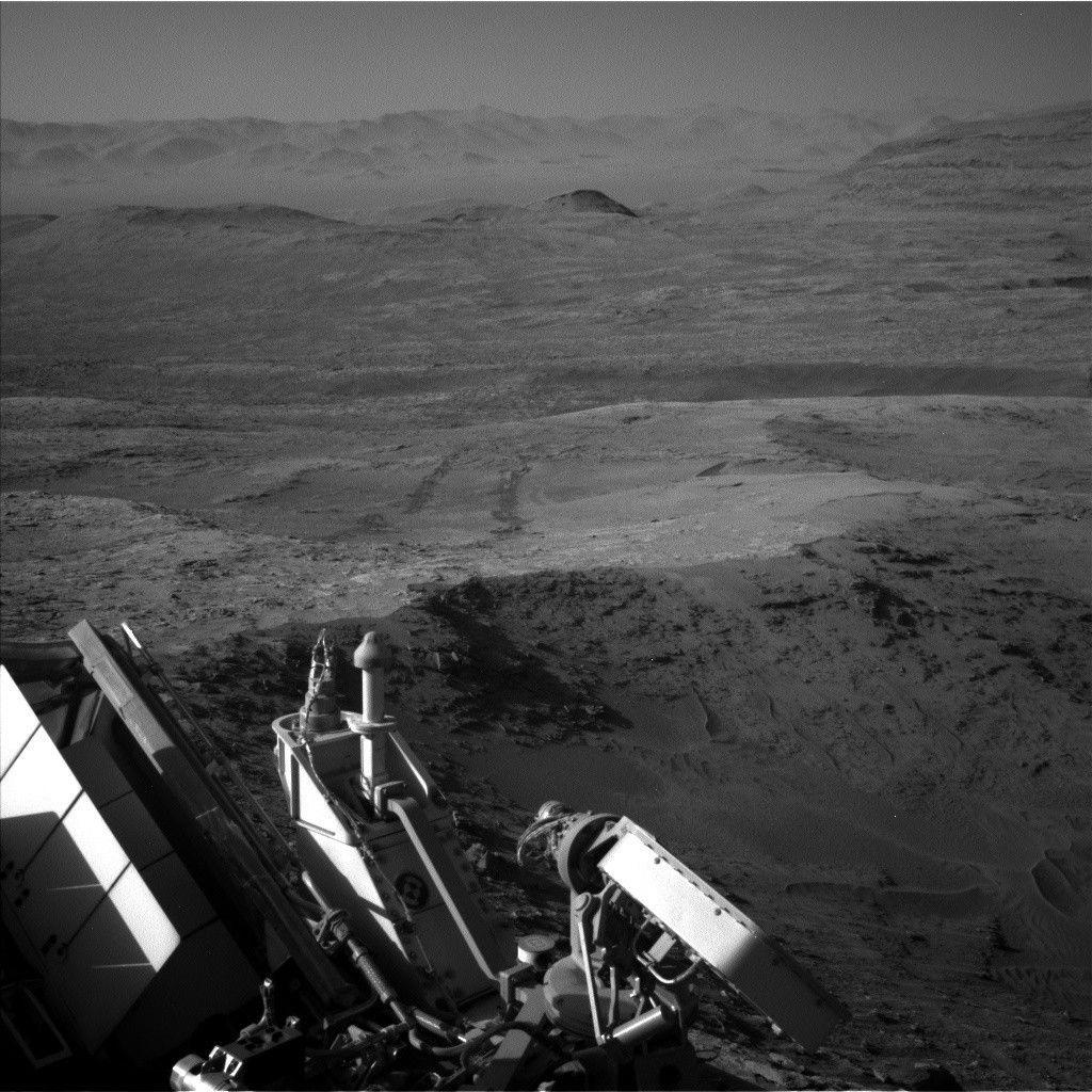 https mars.nasa .gov msl raw images proj msl redops ods surface sol 04612 opgs edr ncam nlb 8069304.jpeg A grayscale photo from the Martian surface shows a landscape of dark gray, slightly uneven, desert-like terrain stretching off into the distance, where a line of peaks that look like sand dunes create a hazy horizon line. Part of the rover is dimly visible in the bottom of the frame, and a pair of wheel tracks, darker than the surrounding ground, extend backward from there, scratched into the soil at the center of the frame.