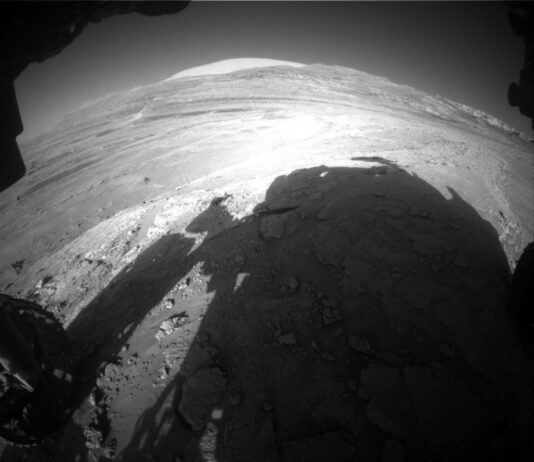 NASA Science: Curiosity Blog Explores Ridge on Sols 4616-4617 An extreme wide-angle, grayscale photo from the Mars surface shows uneven terrain ahead of the rover, covered in numerous small to medium-sized rocks, all light gray and illuminated by sunlight. Impressions in the middle ground ahead form swooping backward Cs, and in the far distance a mountain rises from the ground, but looks very broad and squat, because of the distortion from the wide-angle lens. Parts of the rover are visible around the sides and bottom of the image, and its shadow fills much of the foreground.