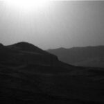 NASA Explores Intriguing Boxwork Formations on Mars’ Surface A grayscale photo from the Martian surface shows a backlit, hilly landscape. In the foreground a pair of peaks on the left side of the image — shadowed and nearly black — descend gently toward the right side, where the ground rises again slightly at the edge of the frame. Beyond that on the right side, a ridge of very dark gray terrain is topped by a wavy line of small peaks, creating a horizon line that disappears behind the foreground hills. The sky is a uniform lighter gray, with a blinding sun visible at the top center of the frame.