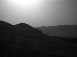 NASA Explores Intriguing Boxwork Formations on Mars’ Surface A grayscale photo from the Martian surface shows a backlit, hilly landscape. In the foreground a pair of peaks on the left side of the image — shadowed and nearly black — descend gently toward the right side, where the ground rises again slightly at the edge of the frame. Beyond that on the right side, a ridge of very dark gray terrain is topped by a wavy line of small peaks, creating a horizon line that disappears behind the foreground hills. The sky is a uniform lighter gray, with a blinding sun visible at the top center of the frame.