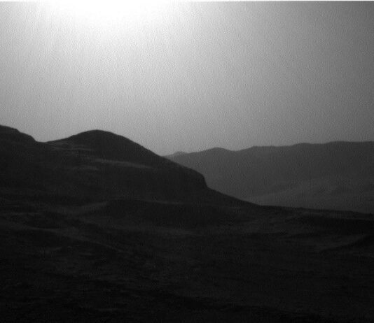NASA Explores Intriguing Boxwork Formations on Mars’ Surface A grayscale photo from the Martian surface shows a backlit, hilly landscape. In the foreground a pair of peaks on the left side of the image — shadowed and nearly black — descend gently toward the right side, where the ground rises again slightly at the edge of the frame. Beyond that on the right side, a ridge of very dark gray terrain is topped by a wavy line of small peaks, creating a horizon line that disappears behind the foreground hills. The sky is a uniform lighter gray, with a blinding sun visible at the top center of the frame.
