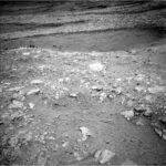 NASA Science: Exploring Hollows on Mars, Sols 4661-4667 A grayscale image from the Martian surface shows dark gray, very rocky terrain. In the foreground, whitish rocks are surrounded by gray soil, while in the middle of the frame a smoother area looks like sand, and beyond that the terrain turns rocky again, sloping downward from the upper right corner of the frame to the upper left side of the image.