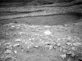 NASA Science: Exploring Hollows on Mars, Sols 4661-4667 A grayscale image from the Martian surface shows dark gray, very rocky terrain. In the foreground, whitish rocks are surrounded by gray soil, while in the middle of the frame a smoother area looks like sand, and beyond that the terrain turns rocky again, sloping downward from the upper right corner of the frame to the upper left side of the image.
