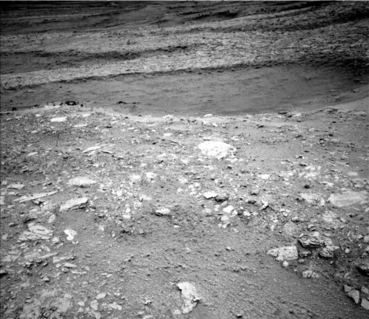 NASA Science: Exploring Hollows on Mars, Sols 4661-4667 A grayscale image from the Martian surface shows dark gray, very rocky terrain. In the foreground, whitish rocks are surrounded by gray soil, while in the middle of the frame a smoother area looks like sand, and beyond that the terrain turns rocky again, sloping downward from the upper right corner of the frame to the upper left side of the image.