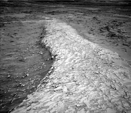 NASA Curiosity Blog: Navigating Sols 4668-4674 Exploration Update A grayscale photo from the Martian surface shows a landscape with an elevated ridge running from the foreground, at the bottom center of the image, weaving off into the distance near the top of the frame. The ridge itself is very light gray and uneven, composed of layered, chipped rock that looks like a dried-out mud flat. Descending off to either side of the ridge, the surrounding terrain is much darker gray and smoother, almost sandy, with scattered small rocks poking above the surface.
