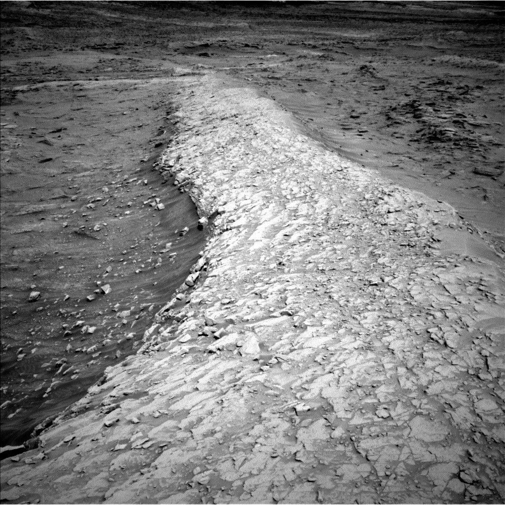 A grayscale photo from the Martian surface shows a landscape with an elevated ridge running from the foreground, at the bottom center of the image, weaving off into the distance near the top of the frame. The ridge itself is very light gray and uneven, composed of layered, chipped rock that looks like a dried-out mud flat. Descending off to either side of the ridge, the surrounding terrain is much darker gray and smoother, almost sandy, with scattered small rocks poking above the surface.