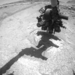 Curiosity Blog: Earthly Spooks, Sols 4702-4708 – NASA Update A wide-angle, grayscale photo from the Mars surface shows mostly flat, medium gray, slightly rough terrain ahead of the rover, with a lighter-toned indentation in the round directly in front of it. In the distance at the upper right of the image, the ground becomes much more uneven and rocky, with a mesa rising from the ground on the horizon beyond that. Parts of the rover are visible, but dark and shadowed. Rover tracks are also visible, zig-zagging on the ground along the left edge of the image. The end of the rover’s robotic arm, with its fist-like collection of instruments, is poised just above the lighter-colored indentation in the ground, and its shadow runs on the ground back to the rover body at the bottom of the frame.