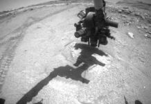 Curiosity Blog: Earthly Spooks, Sols 4702-4708 – NASA Update A wide-angle, grayscale photo from the Mars surface shows mostly flat, medium gray, slightly rough terrain ahead of the rover, with a lighter-toned indentation in the round directly in front of it. In the distance at the upper right of the image, the ground becomes much more uneven and rocky, with a mesa rising from the ground on the horizon beyond that. Parts of the rover are visible, but dark and shadowed. Rover tracks are also visible, zig-zagging on the ground along the left edge of the image. The end of the rover’s robotic arm, with its fist-like collection of instruments, is poised just above the lighter-colored indentation in the ground, and its shadow runs on the ground back to the rover body at the bottom of the frame.