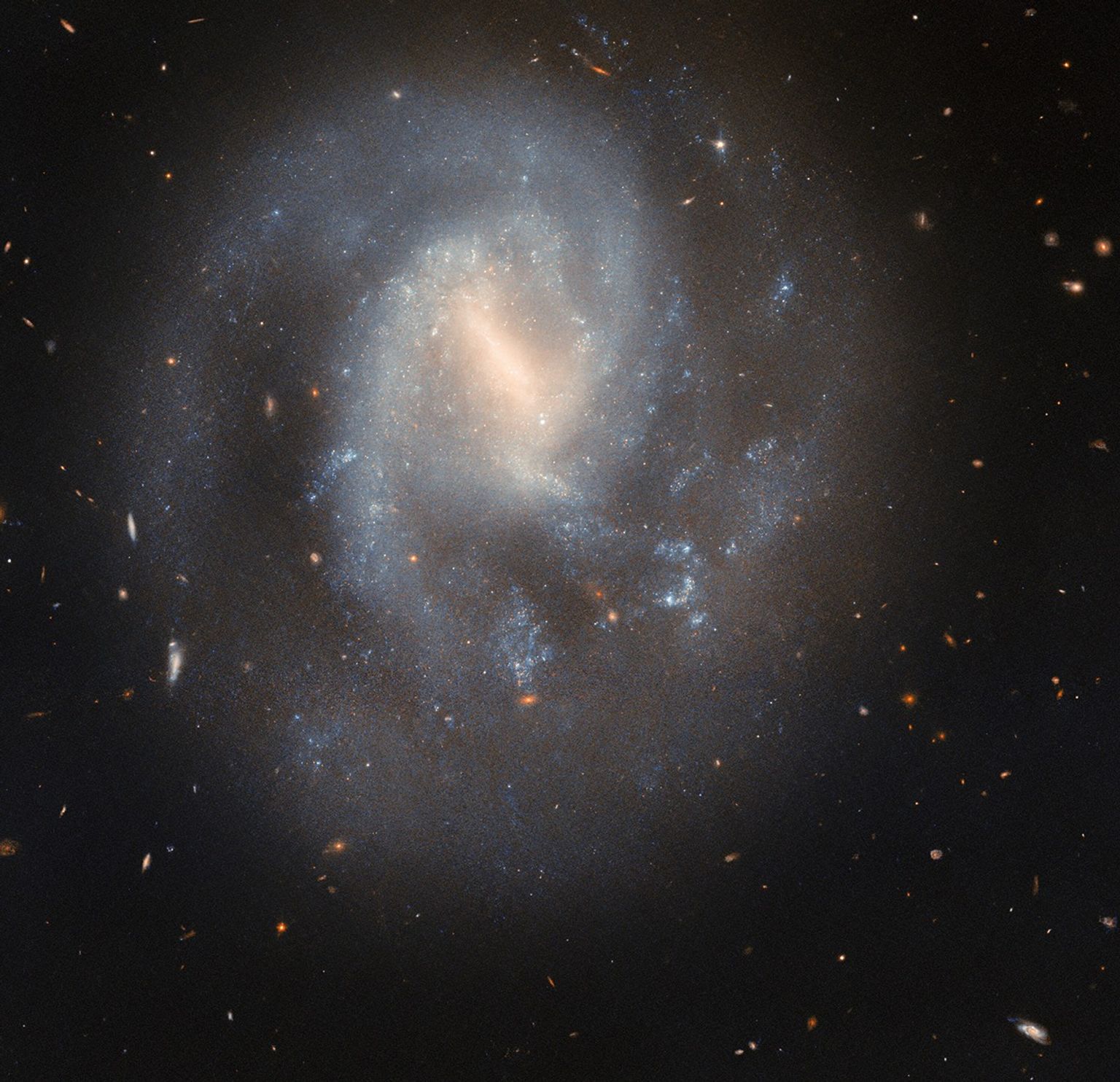 A spiral galaxy with a soft and slightly faint appearance. It glows most brightly around the pale-yellow bar across its center. It has two spiral arms which wrap around its core, quickly broadening out to join a wide, faint circular halo around the galaxy. Glowing, sparkling patches in the disk show stars forming in nebulae. Behind the galaxy, distant galaxies appear as orange dots on a black background.
